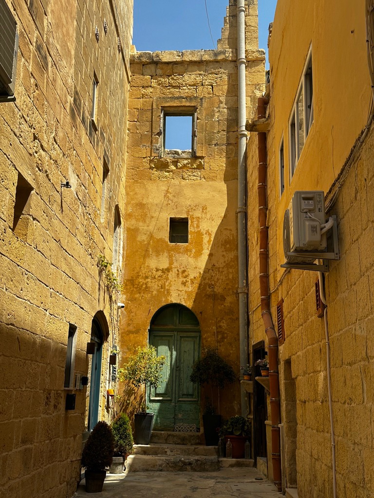 A narrow yellow limestone alley in Gozo with a green arched door and a window opening to the sky